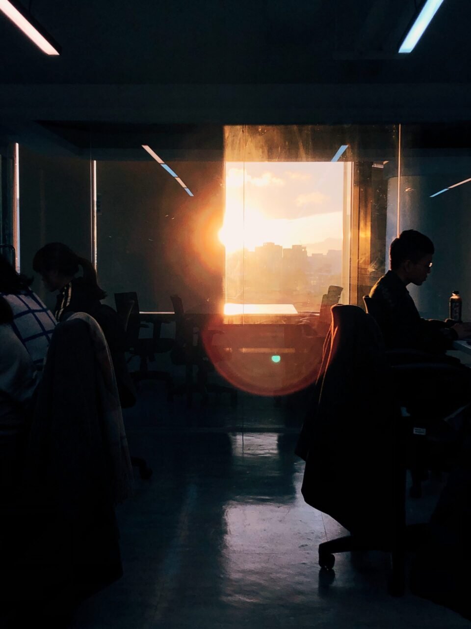 Office workers silhouetted by a bright sunset shining through the large window.