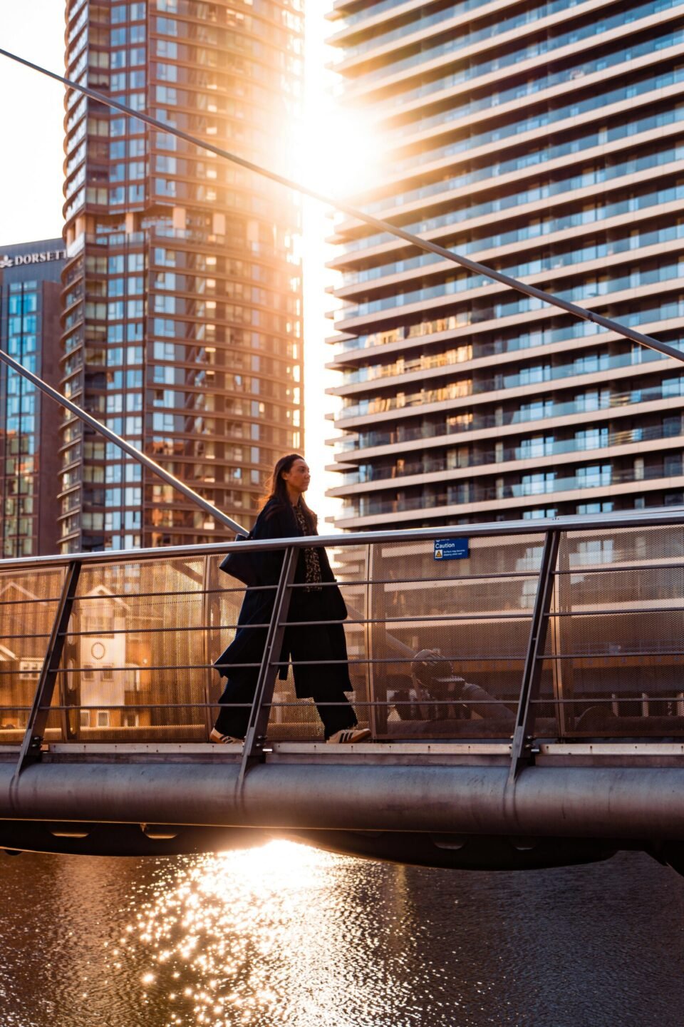 Person walking across a modern bridge in a city, backlit by bright sunset and tall glass buildings.